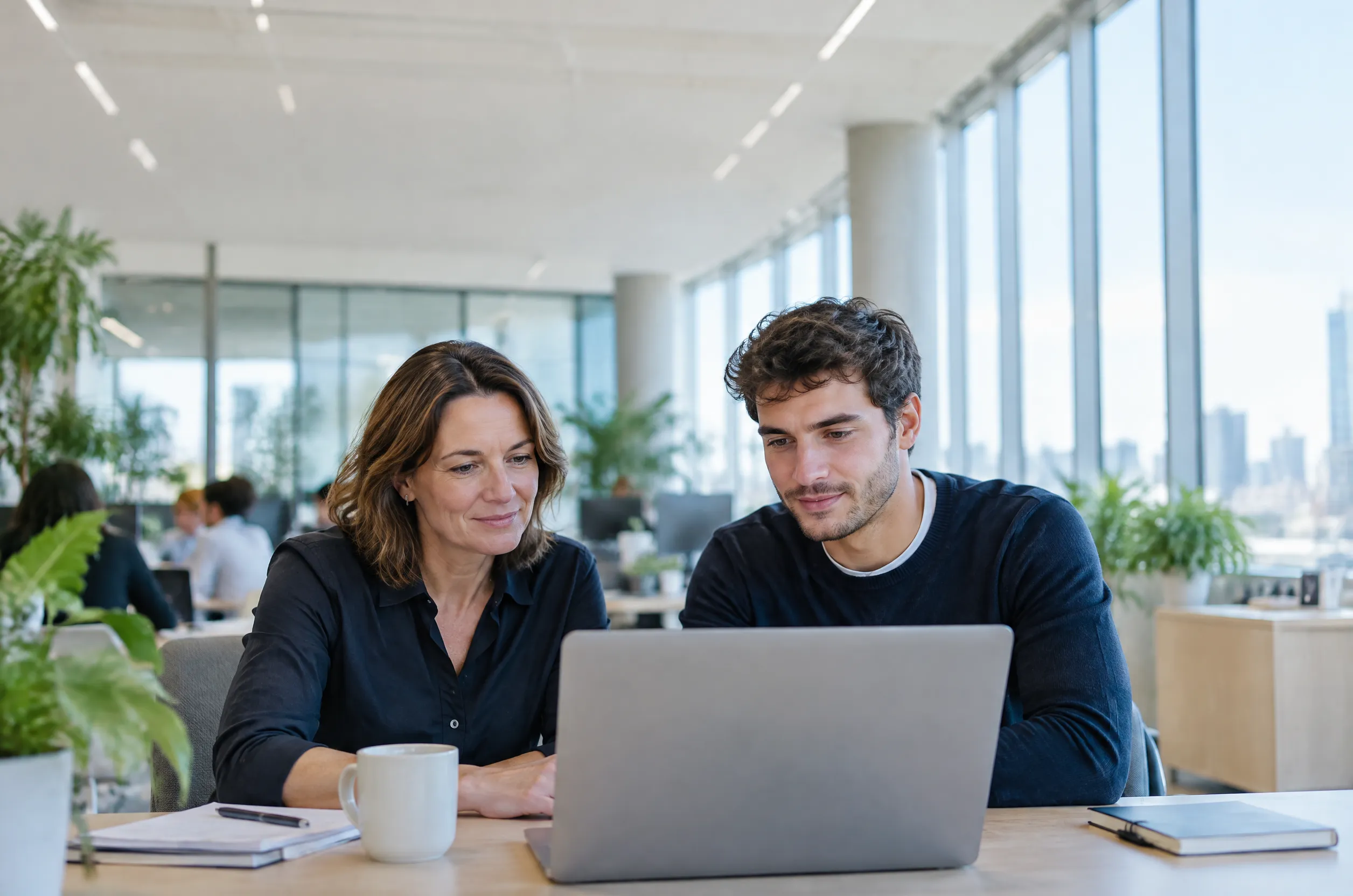 Two colleagues review a laptop screen together at a desk in a bright open-plan office.